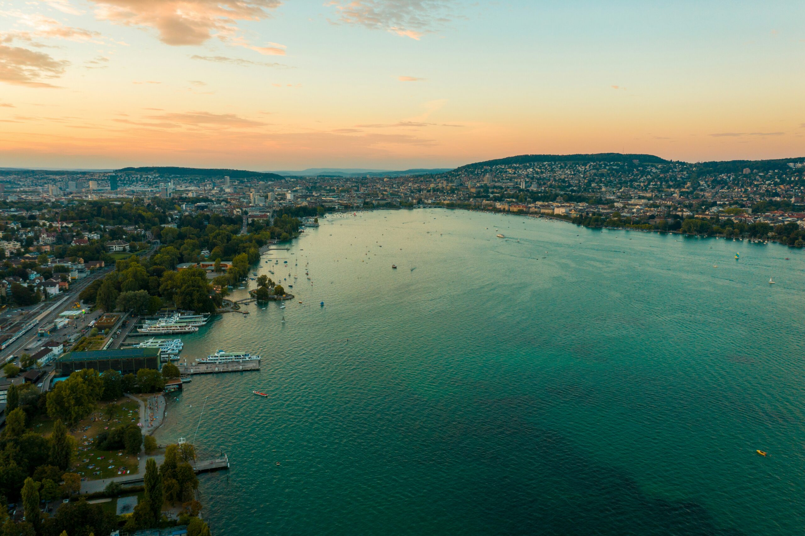 A scenic, wide-angle shot of Lake Zurich under a partly cloudy sky. The calm, blue water stretches out towards distant hills and mountains on the horizon. On the right, a lush green shoreline is visible with trees and buildings, while a few small white boats can be seen sailing on the lake. The water near the foreground is clear, revealing stones on the lakebed.