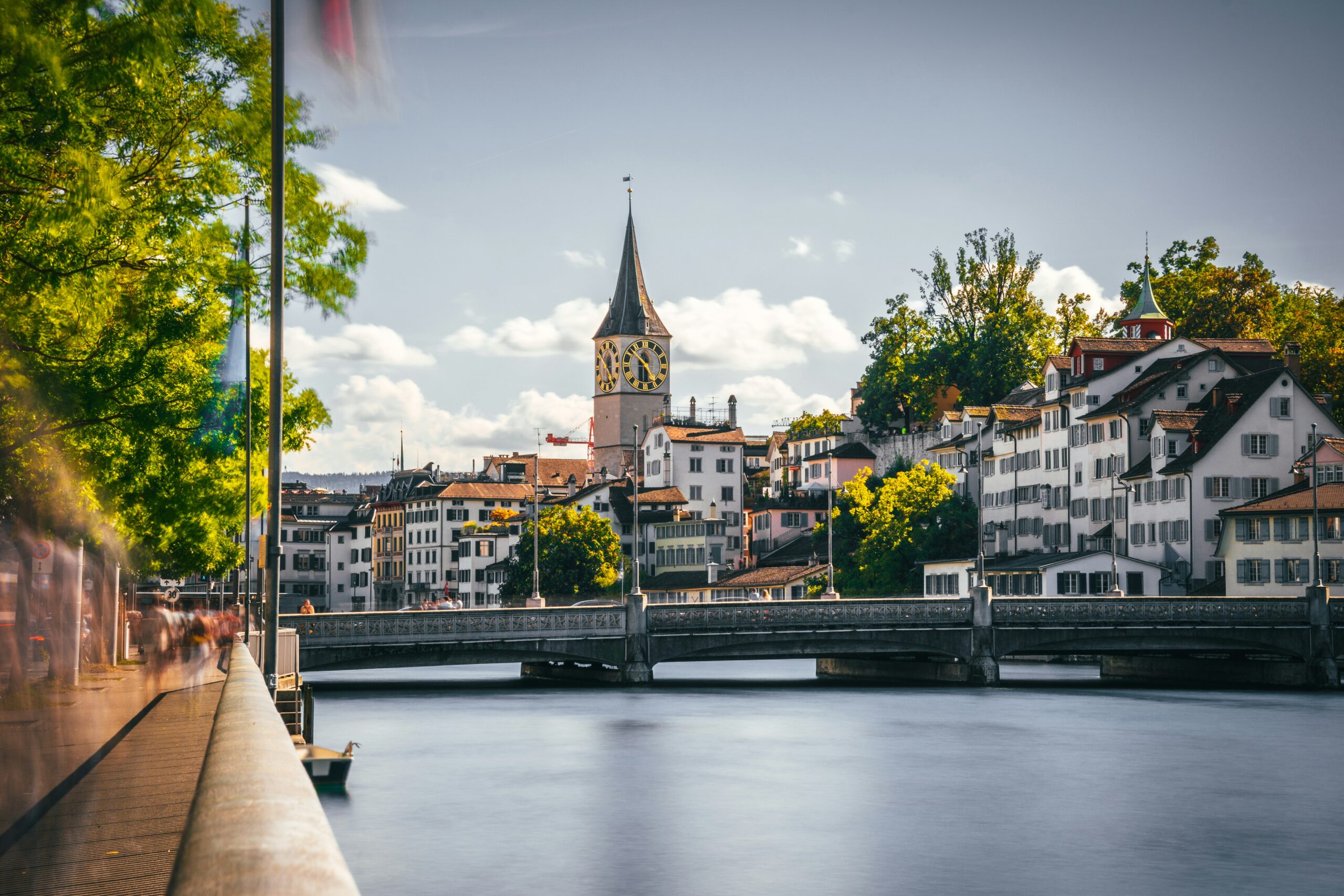 A picturesque view of Zurich's Old Town (Altstadt) centered on the Limmat River. The calm water reflects the historic architecture lining the riverbanks. On the right stands the iconic Grossmünster church with its twin towers, while the left bank features the slender teal spire of the Fraumünster church. The scene is framed by a clear blue sky and connects the two sides of the city with a low stone bridge in the distance.
